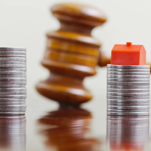 A miniature house balanced on a stack of coins, with a gavel and another stack of coins in the blurred background.